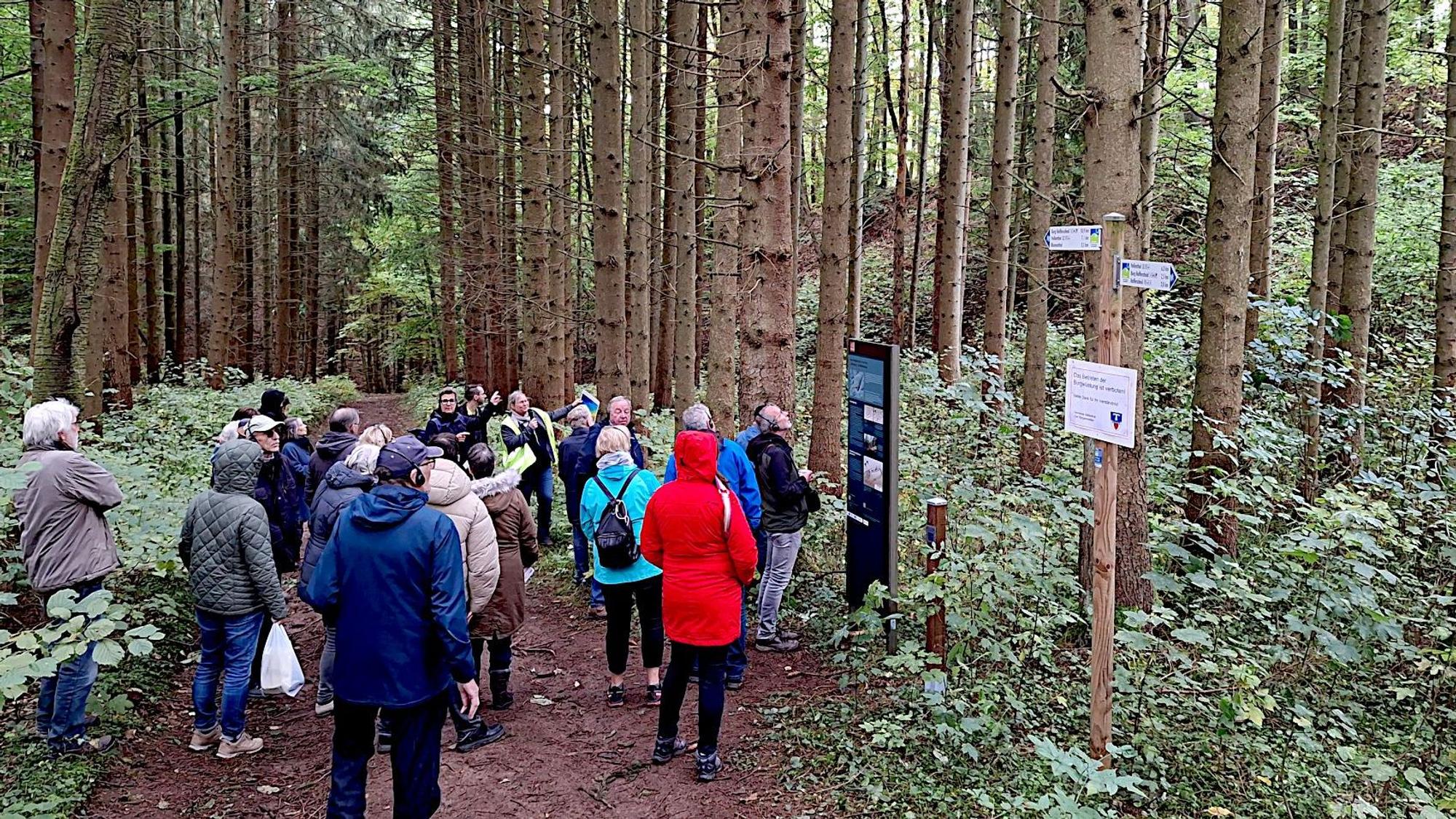 Eine Gruppe von Menschen steht vor einem Hinweisschild an einem Weg, der durch einen Fichtenwald verläuft.