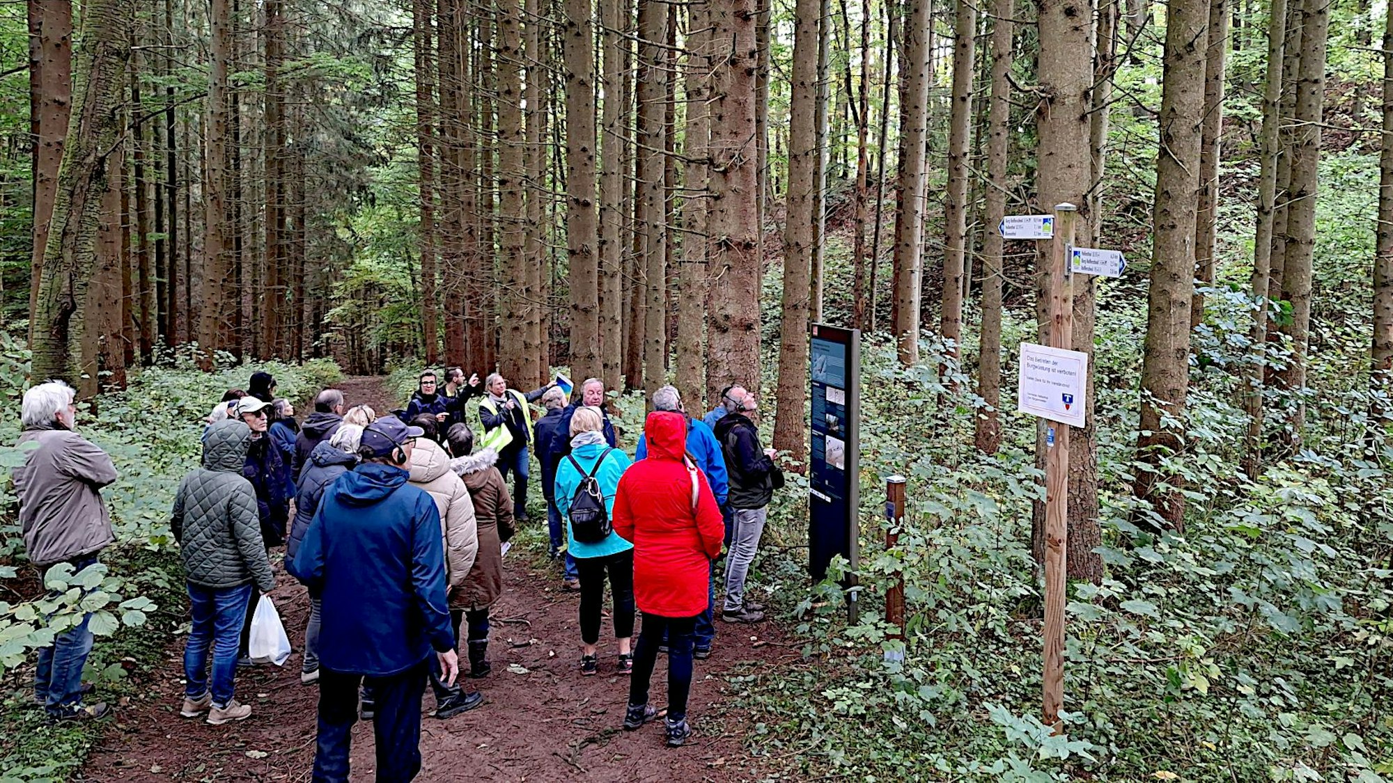 Eine Gruppe von Menschen steht vor einem Hinweisschild an einem Weg, der durch einen Fichtenwald verläuft.
