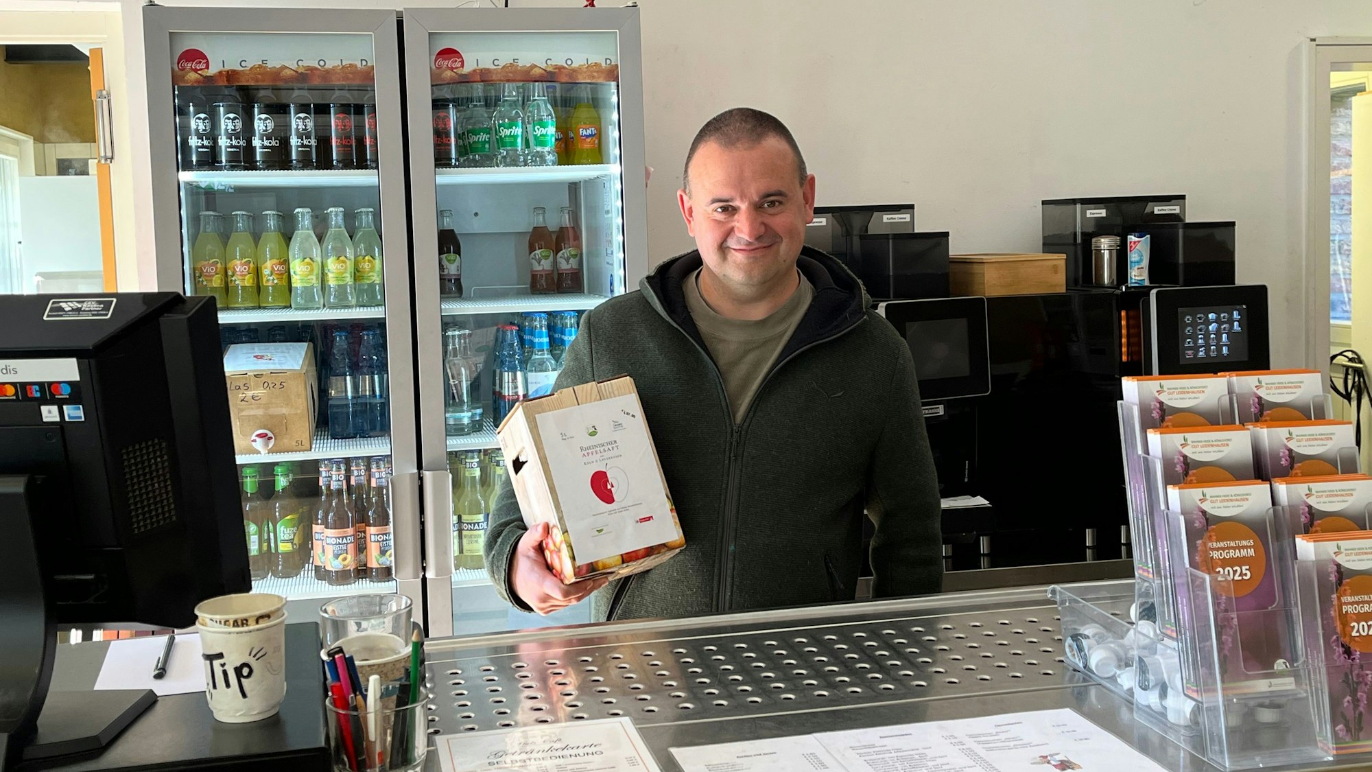 Ein Mann steht hinter einer Verkaufstheke und hat einen Karton Apfelsaft in der Hand.