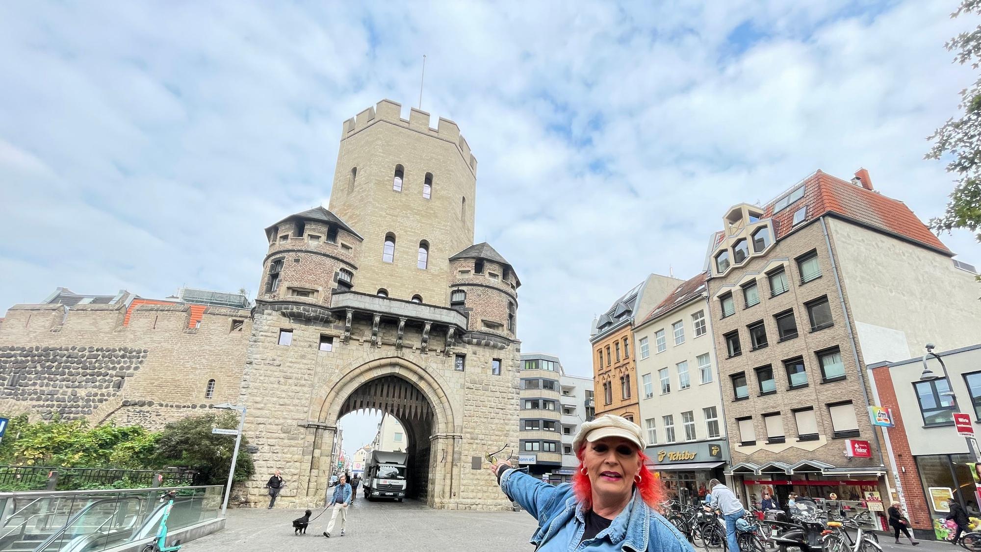 Eine Frau mit roten Haaren und Kappe, die Travestie-Künstlerin Swanee Feels, steht vor der Severinstorburg.