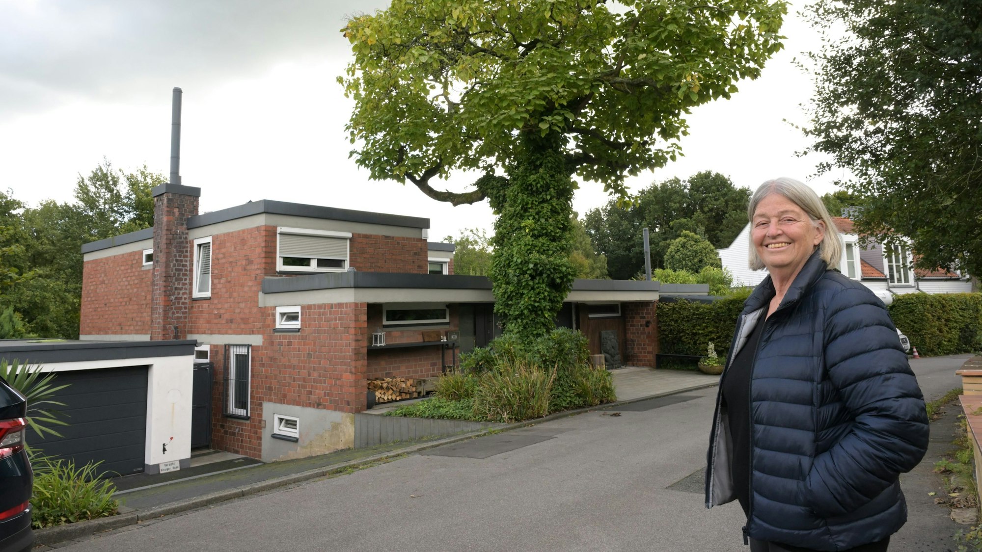 Ein funktionalistischer Klinkerbau mit Flachdach. Davor ein Baum. An der Seite steht eine ältere Frau mit grauer Kurzhaarfrisur.
