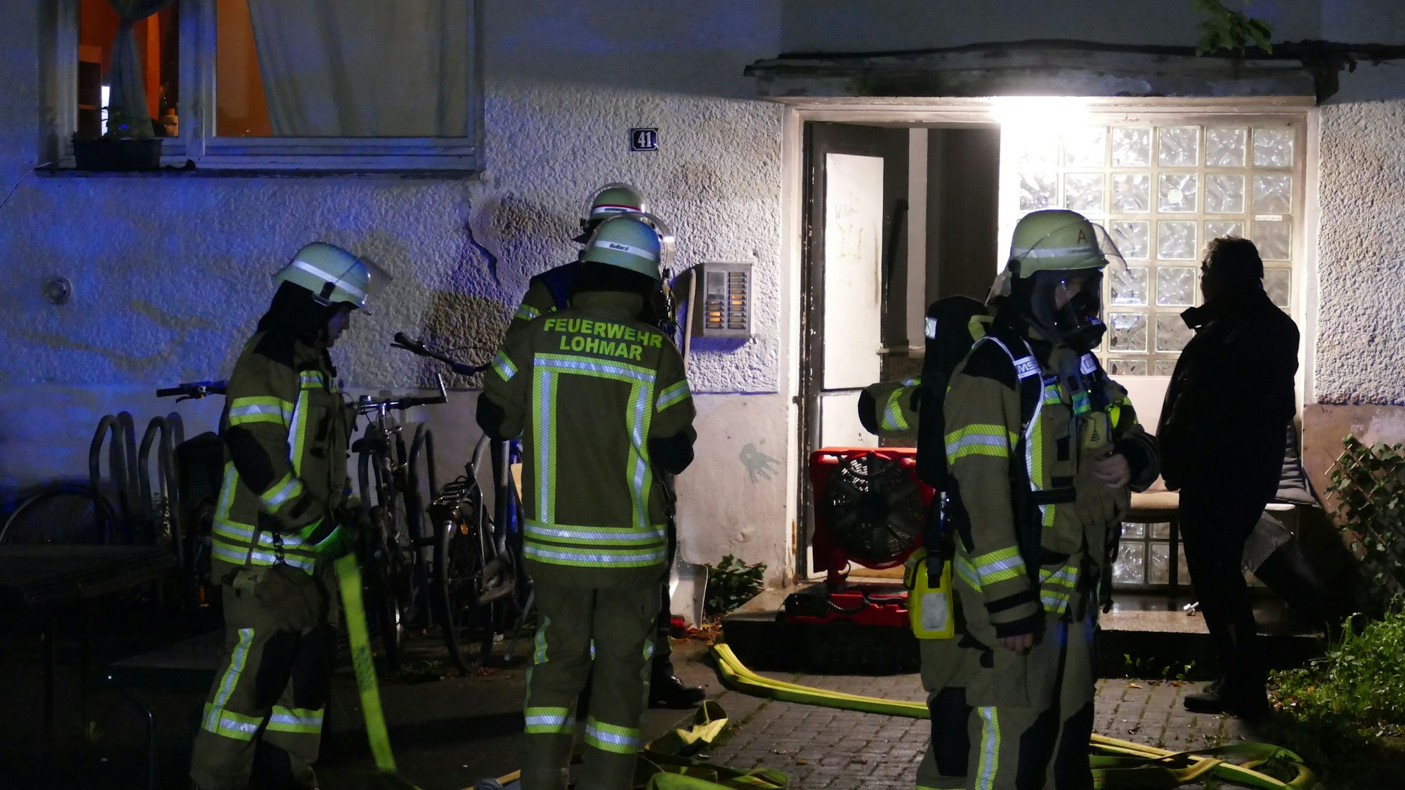 Feuerwehrleute stehen vor dem Eingang der Wohnungslosen-Unterkunft am Dammweg in Lohmar, in der es brannte.