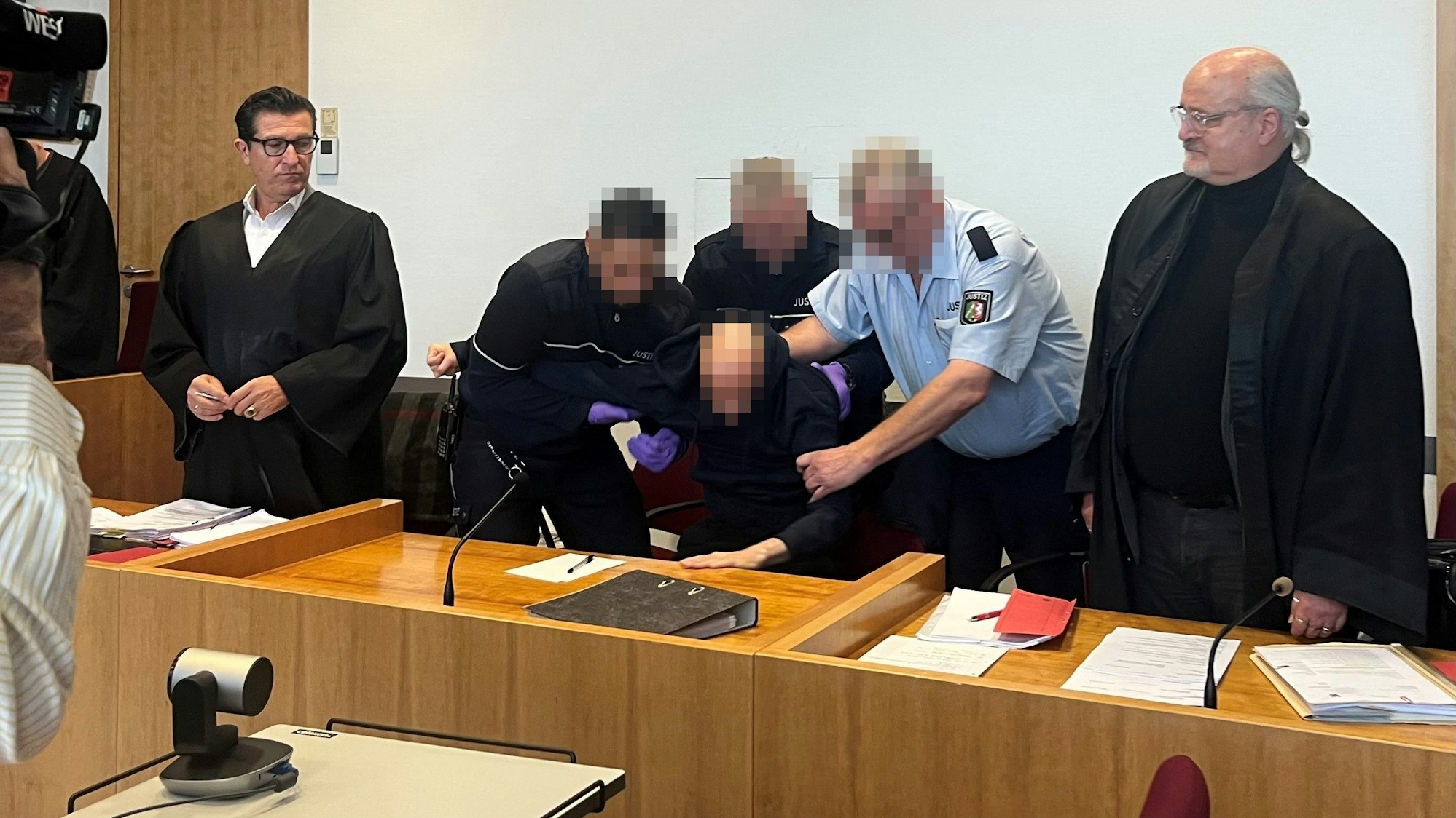 Drei Justizwachtmeister zwingen den Angeklagten, aufzustehen. Er hatte sich geweigert, sich beim Eintreten der Kammer in den Saal zu erheben und das Gericht beleidigt. Links Verteidiger Michael Hakner, rechts Verteidiger Bernd Arnold.