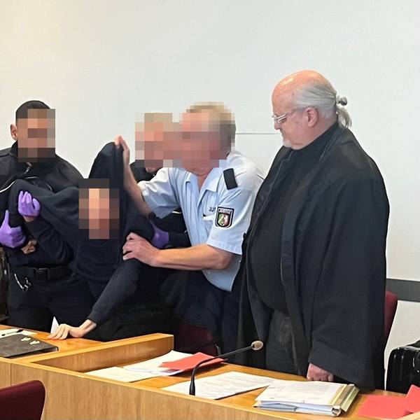Drei Justizwachtmeister zwingen den Angeklagten, aufzustehen. Er hatte sich geweigert, sich beim Eintreten der Kammer in den Saal zu erheben und das Gericht beleidigt. Links Verteidiger Michael Hafner, rechts Verteidiger Bernd Arnold.