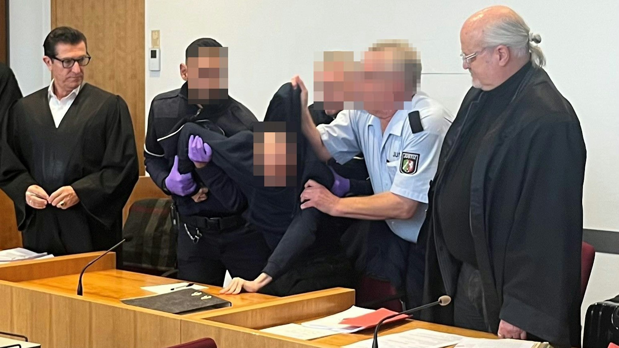 Drei Justizwachtmeister zwingen den Angeklagten, aufzustehen. Er hatte sich geweigert, sich beim Eintreten der Kammer in den Saal zu erheben und das Gericht beleidigt. Links Verteidiger Michael Hafner, rechts Verteidiger Bernd Arnold.