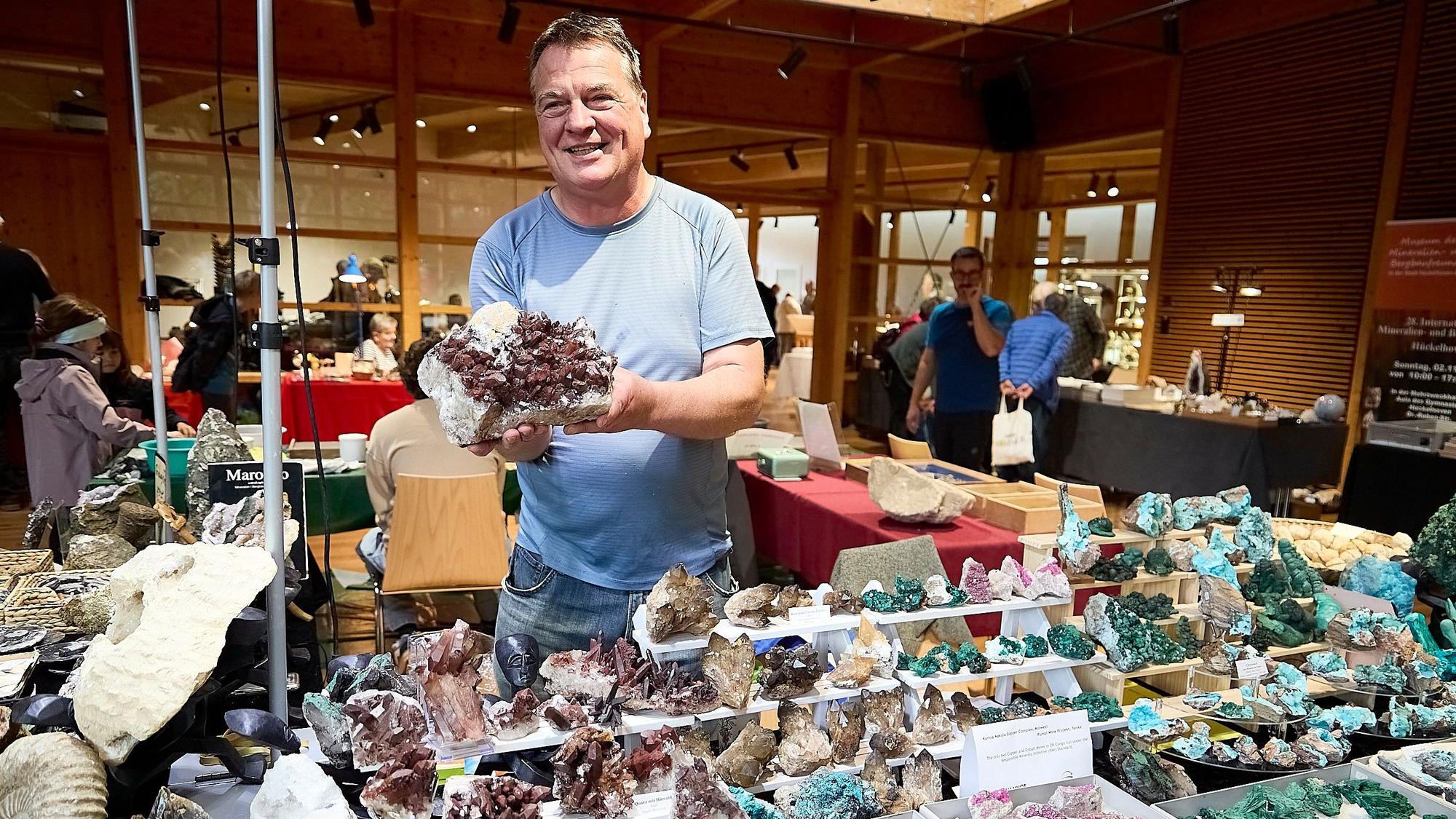 Bernhard Erkelenz hält ein Mineralgestein in die Luft. Vor ihm sind Mineralien und Fossilen ausgestellt.