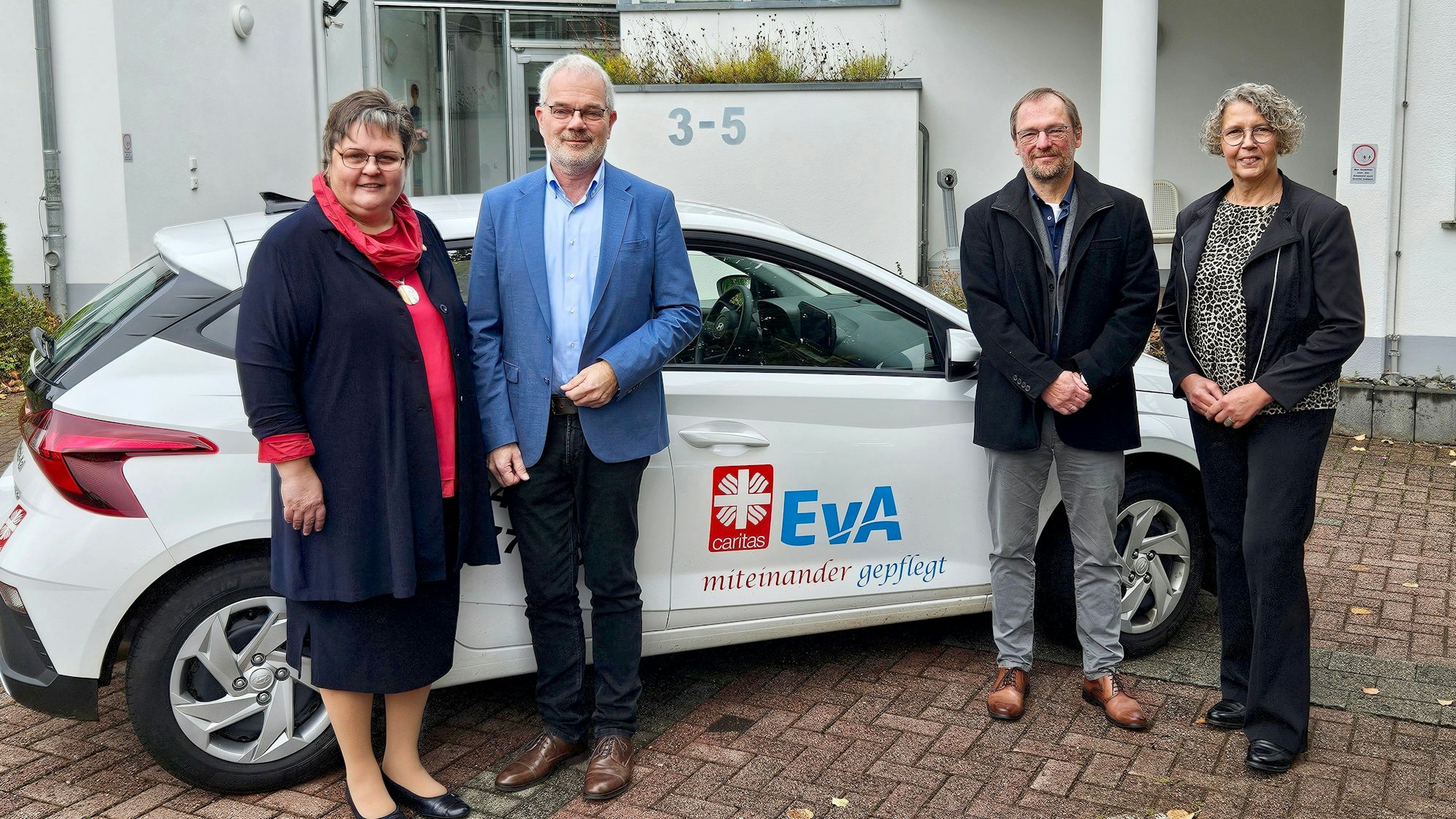 Ute Stolz, Malte Duisberg, Pfarrer Christoph Ude und Elisabeth Nosbers stehen vor einem neuen Wagen, den beide Logos zieren.