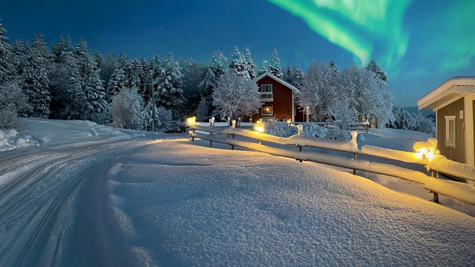 Das Bild zeigt eine Winterlandschaft. Eine schneebedeckte Straße führt an gemütlichen Holzhäusern vorbei. Über der Szene tanzen grüne Nordlichter am klaren Nachthimmel.