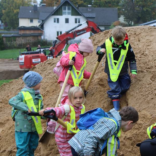 Mehrere Kinder mit Schaufeln auf einem Sandhügel, im Hintergrund ist ein Bagger zu sehen.