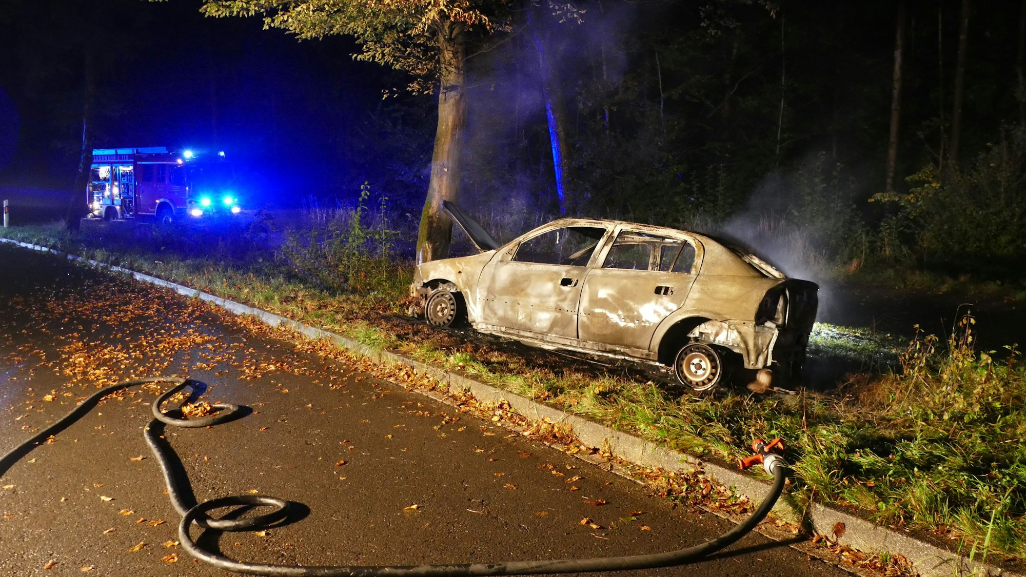 Ein ausgebrannter Wagen steht an einem Baum. In der Luft hängen noch Rauchschwaden, im Hintergrund steht ein Feuerwehrauto mit Blaulicht.