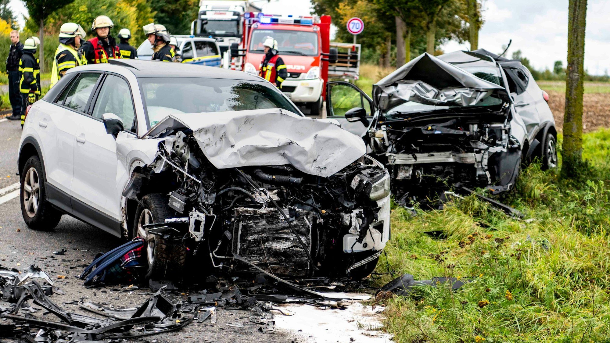 Nach einem Verkehrsunfall stehen zwei stark demolierte Autos auf der Straße, im Hintergrund sind Einsatzkräfte der Feuerwehr zu erkennen.