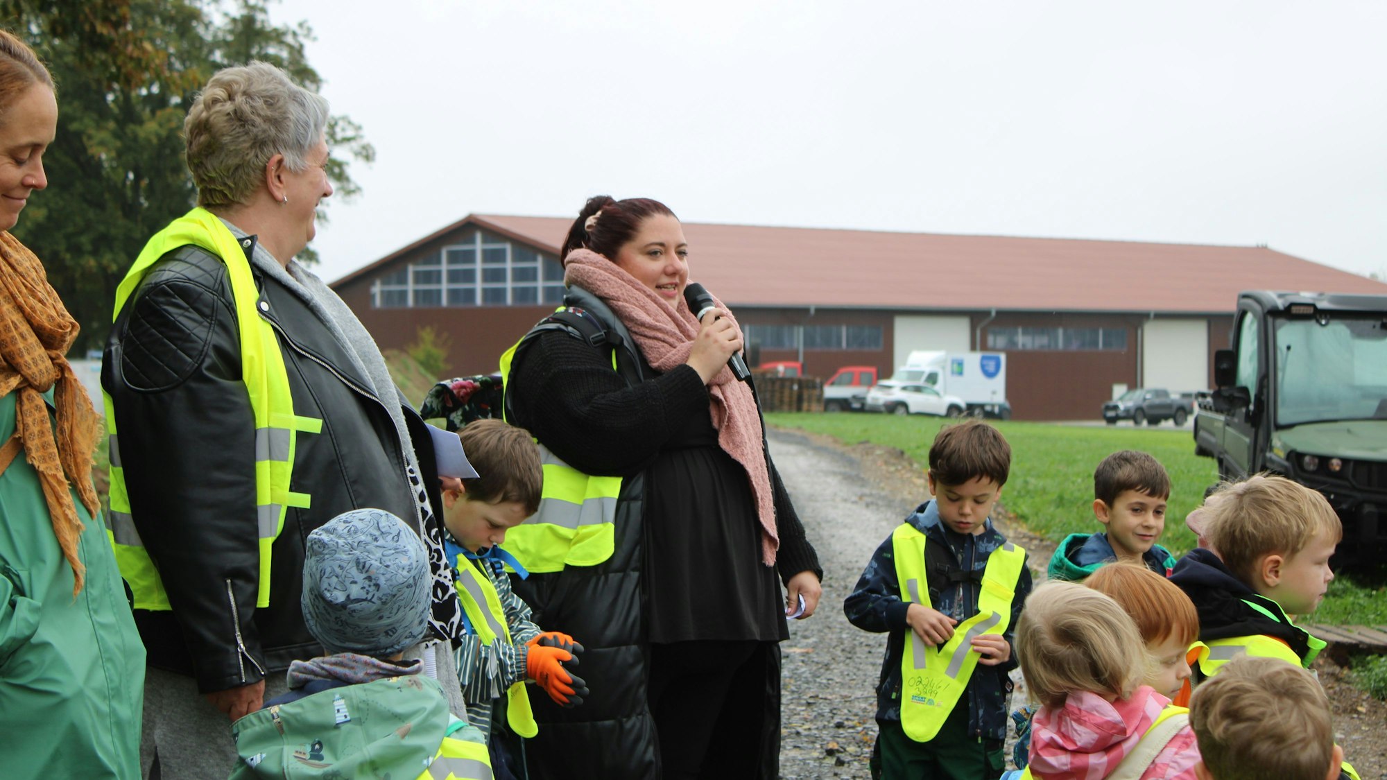 Eine Frau spricht in ein Mikrophon, um sie herum mehrere Kinder, im Hintergrund ist das Gebäude eines Hofs zu sehen.