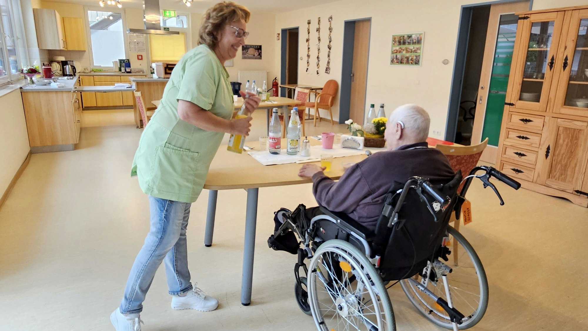 Eine Pflegerin reicht einem Mann, der im Rollstuhl sitzt, etwas zu trinken.