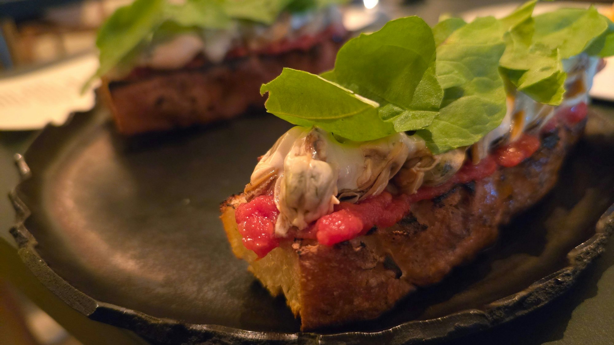 Ein mit Paste und Muscheln belegtes Brot ist zu sehen, große grüne Rauke-Blätter liegen obenauf.