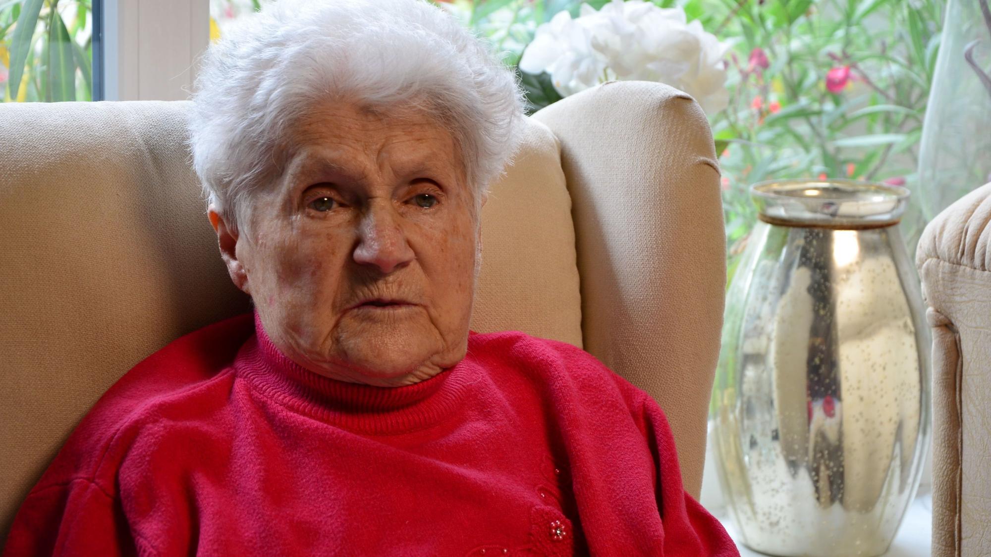 Das Bild zeigt die Dame auf ihrem Sessel. Sie trägt einen roten Pullover.