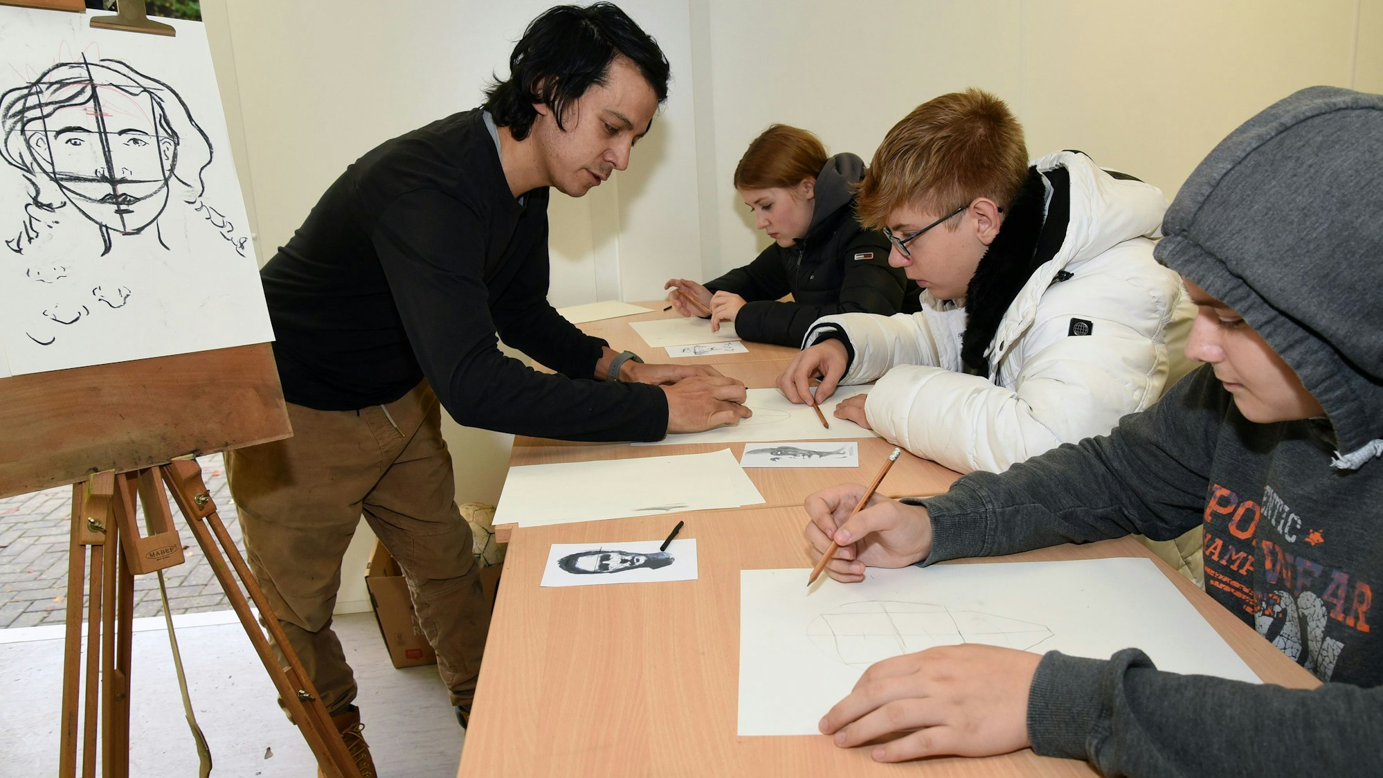 Der Künstler Mauricio Jimenez (links) aus Gummersbach-Hülsenbuch zeigt den Kindern und Jugendlichen in Waldbröl, wie mit Kohlestiften Porträtbilder entstehen.