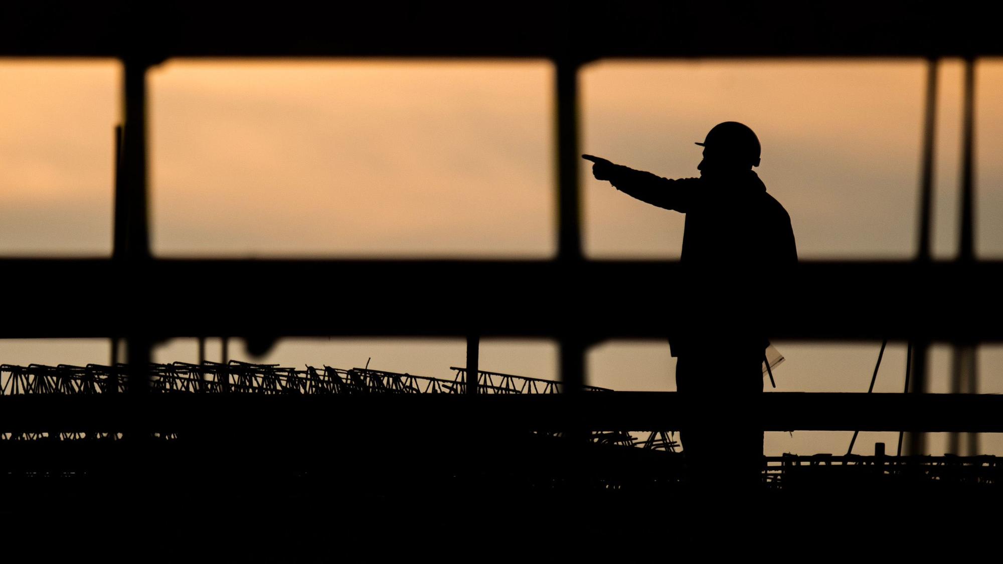 Die Silhouette eines Bauarbeiters zeichnet sich auf einer Baustelle vor dem verfärbten Morgenhimmel ab.