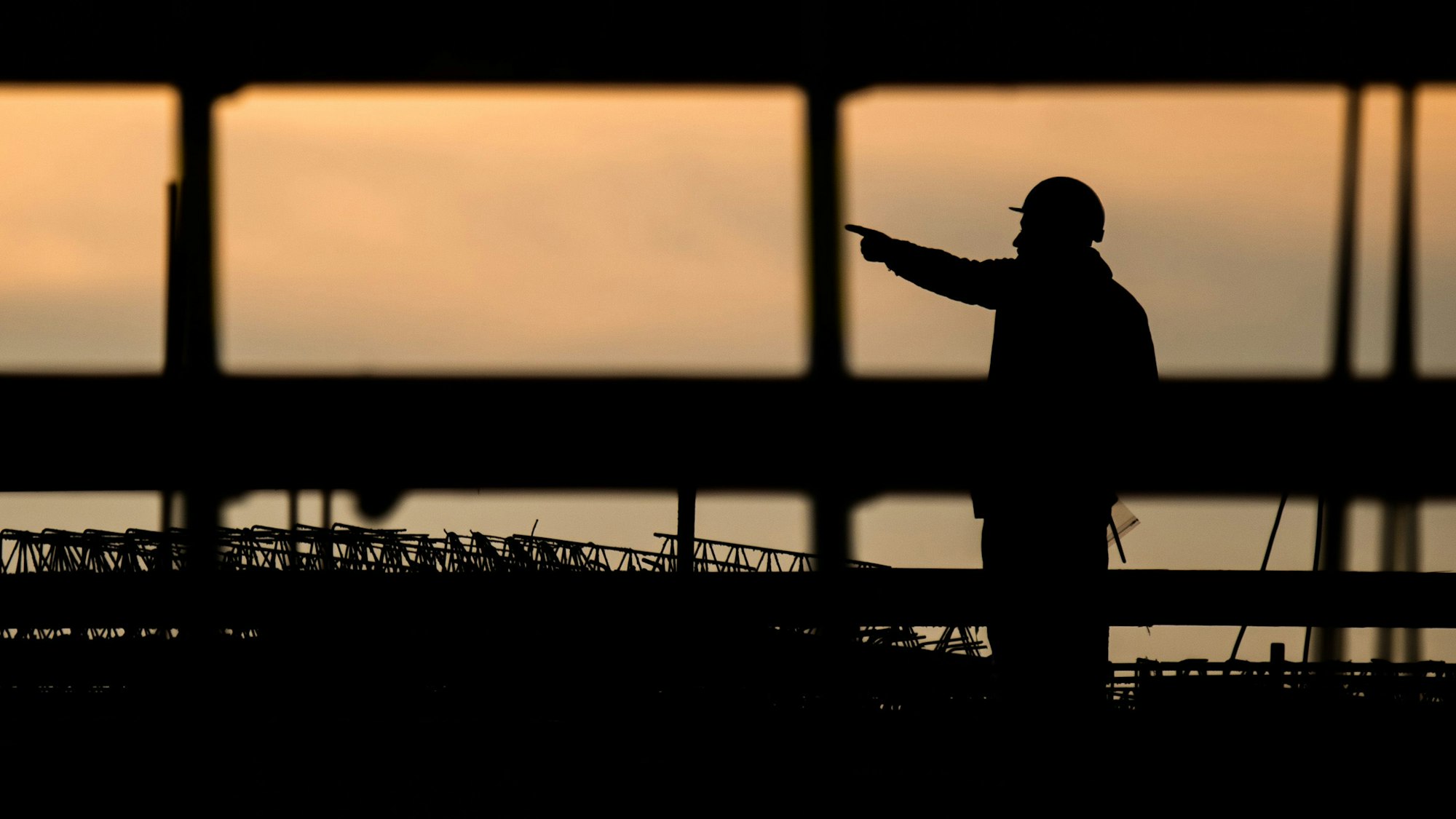 Die Silhouette eines Bauarbeiters zeichnet sich auf einer Baustelle vor dem verfärbten Morgenhimmel ab.