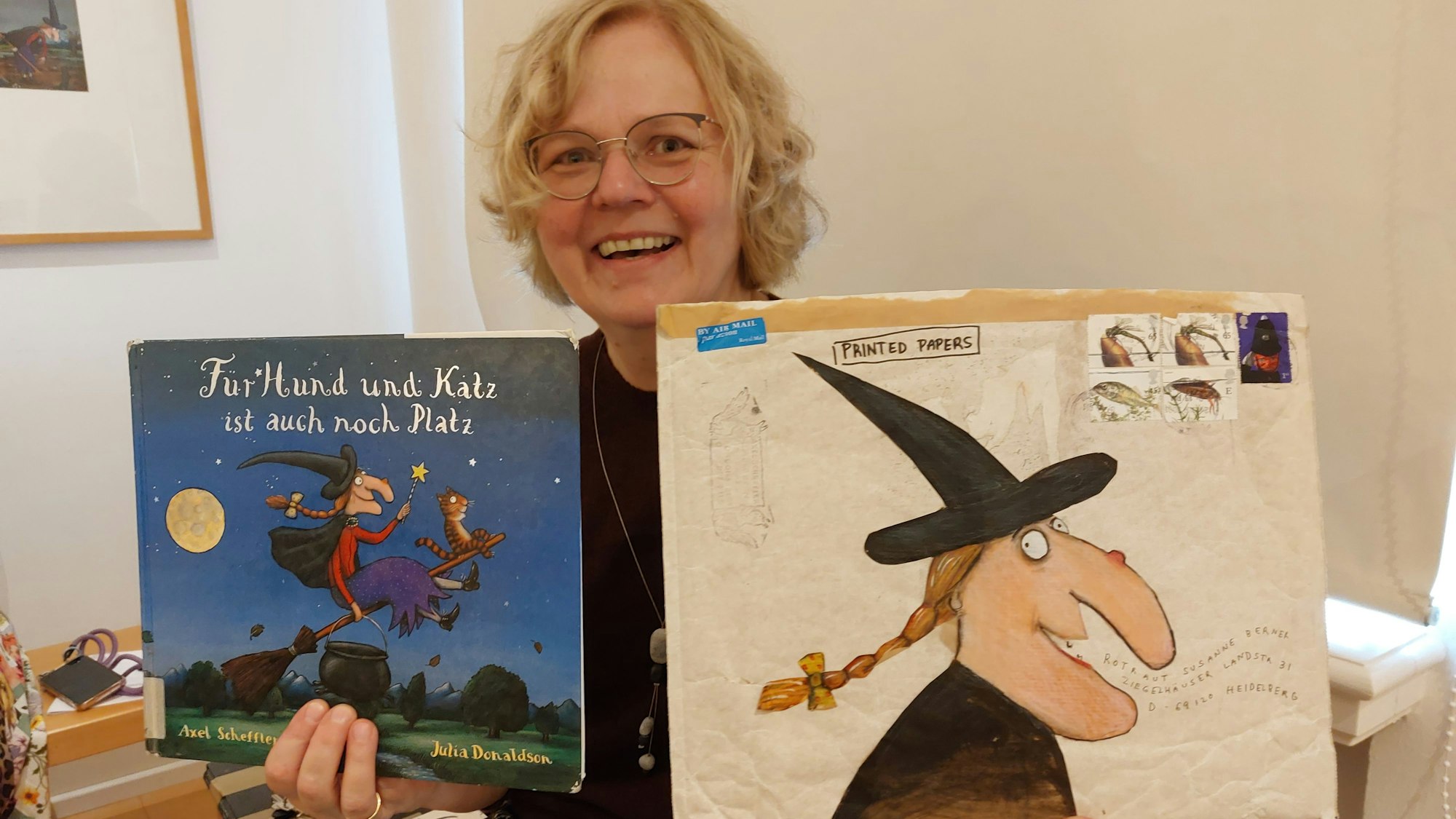 Eine blonde Frau zeigt ein Bilderbuch und die ganz ähnliche Zeichnung einer Hexe auf einem großen Briefumschlag.