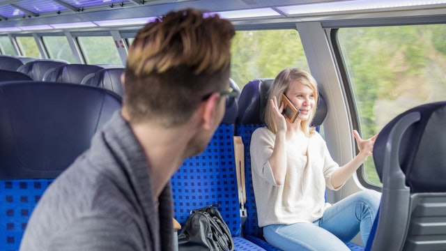In einer Regionalbahn sitzt eine gestikulierende, mit Handy telefonierende junge Frau. Ein junger Mann, der in einer Platzreihe vor ihr sitzt, dreht sich zu ihr um.