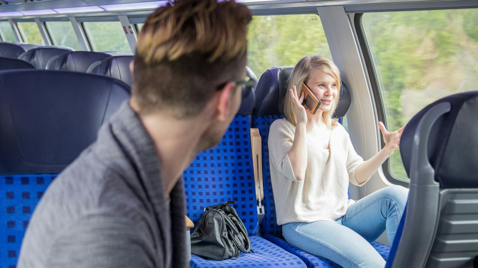 In einer Regionalbahn sitzt eine gestikulierende, mit Handy telefonierende junge Frau. Ein junger Mann, der in einer Platzreihe vor ihr sitzt, dreht sich zu ihr um.