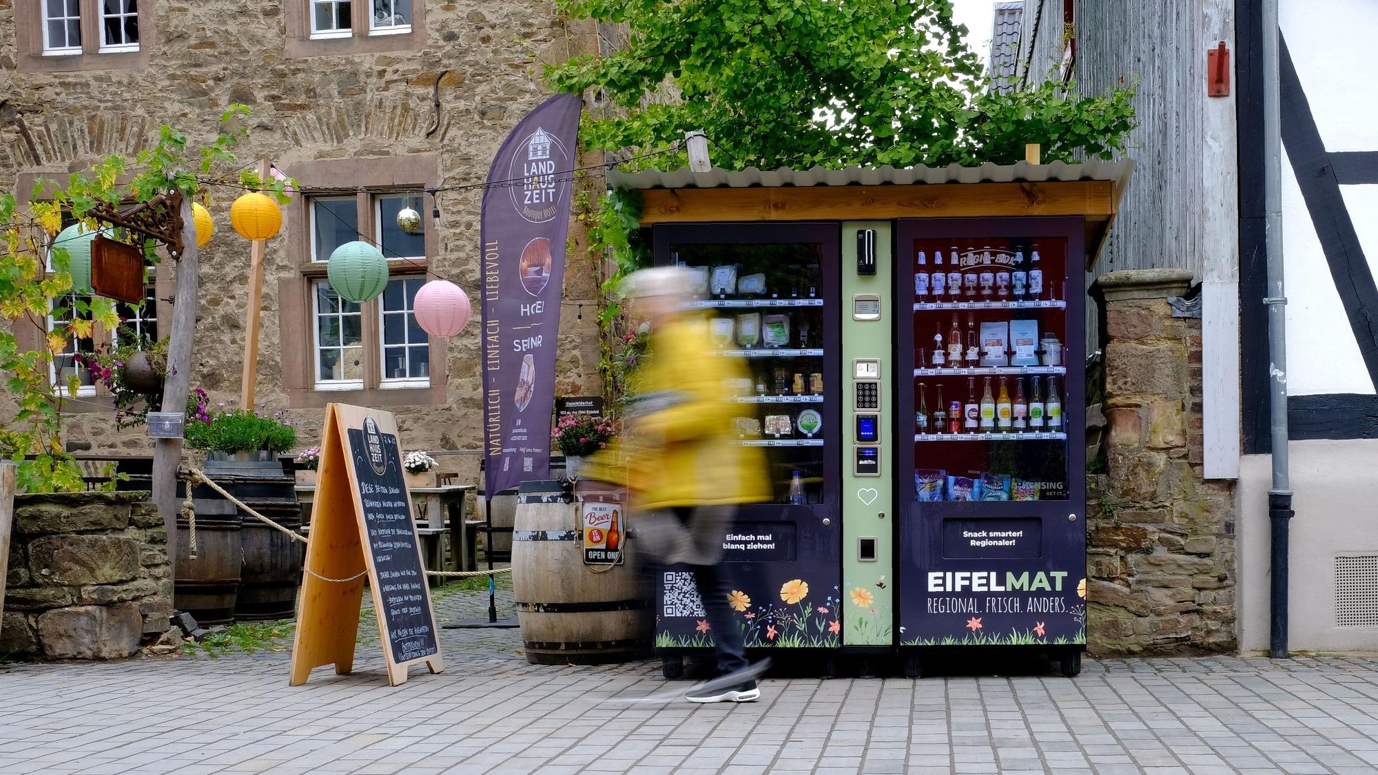 An einem Verkaufsautomaten, der in der Fußgängerzone in Bad Münstereifel steht, gehen Passanten vorbei, die bewegungsunscharf dargestellt werden.