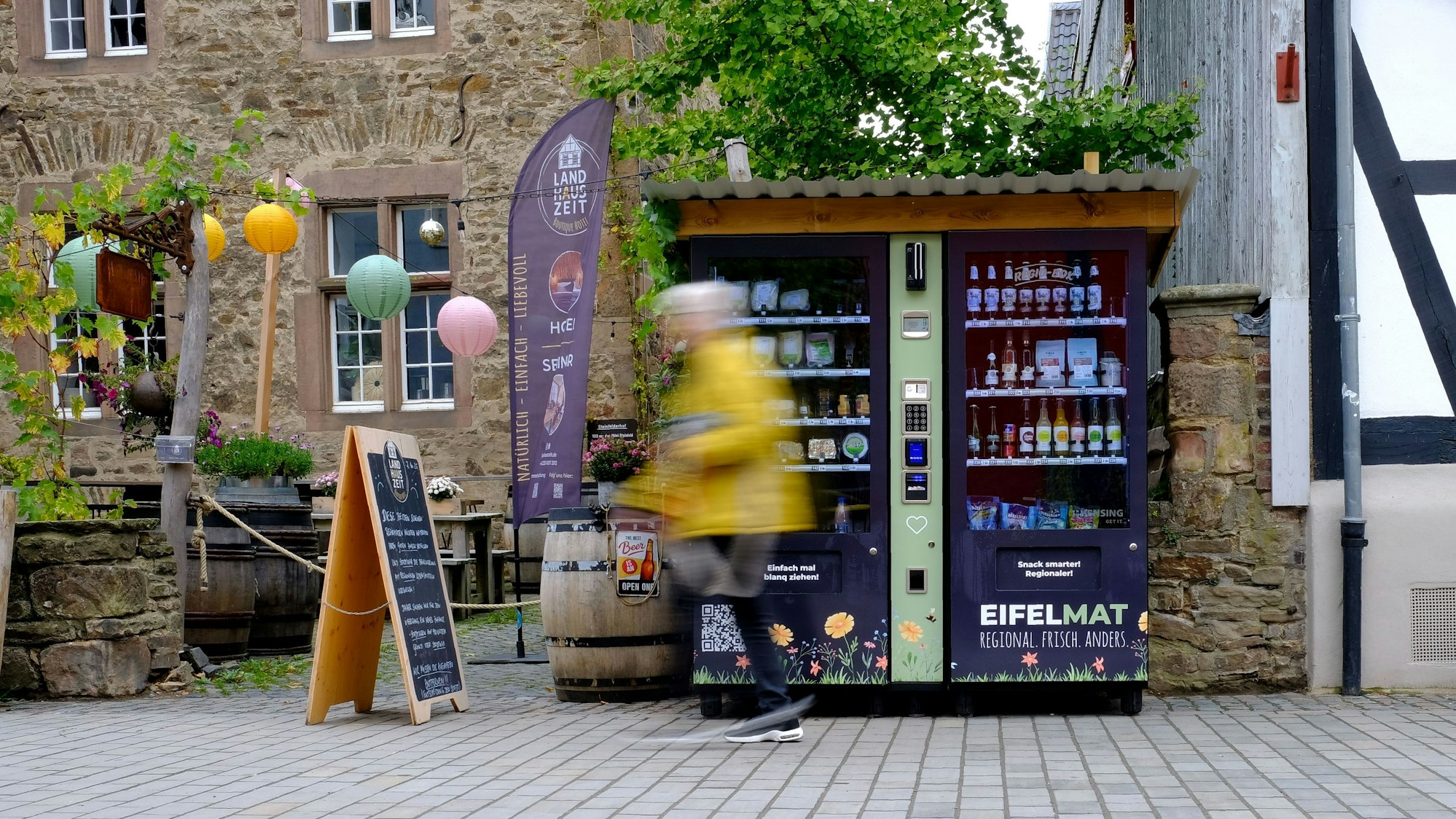 An einem Verkaufsautomaten, der in der Fußgängerzone in Bad Münstereifel steht, gehen Passanten vorbei, die bewegungsunscharf dargestellt werden.