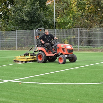 Ein Mann sitzt auf einem kleinen Trecker und zieht damit eine große Bürste über einen Kunstrasenplatz.