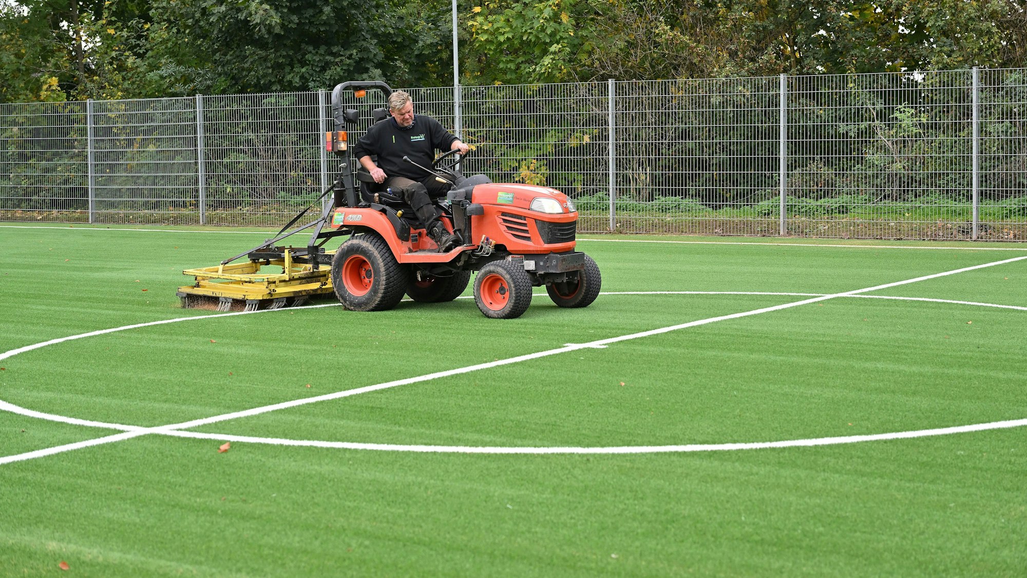 Ein Mann sitzt auf einem kleinen Trecker und zieht damit eine große Bürste über einen Kunstrasenplatz.