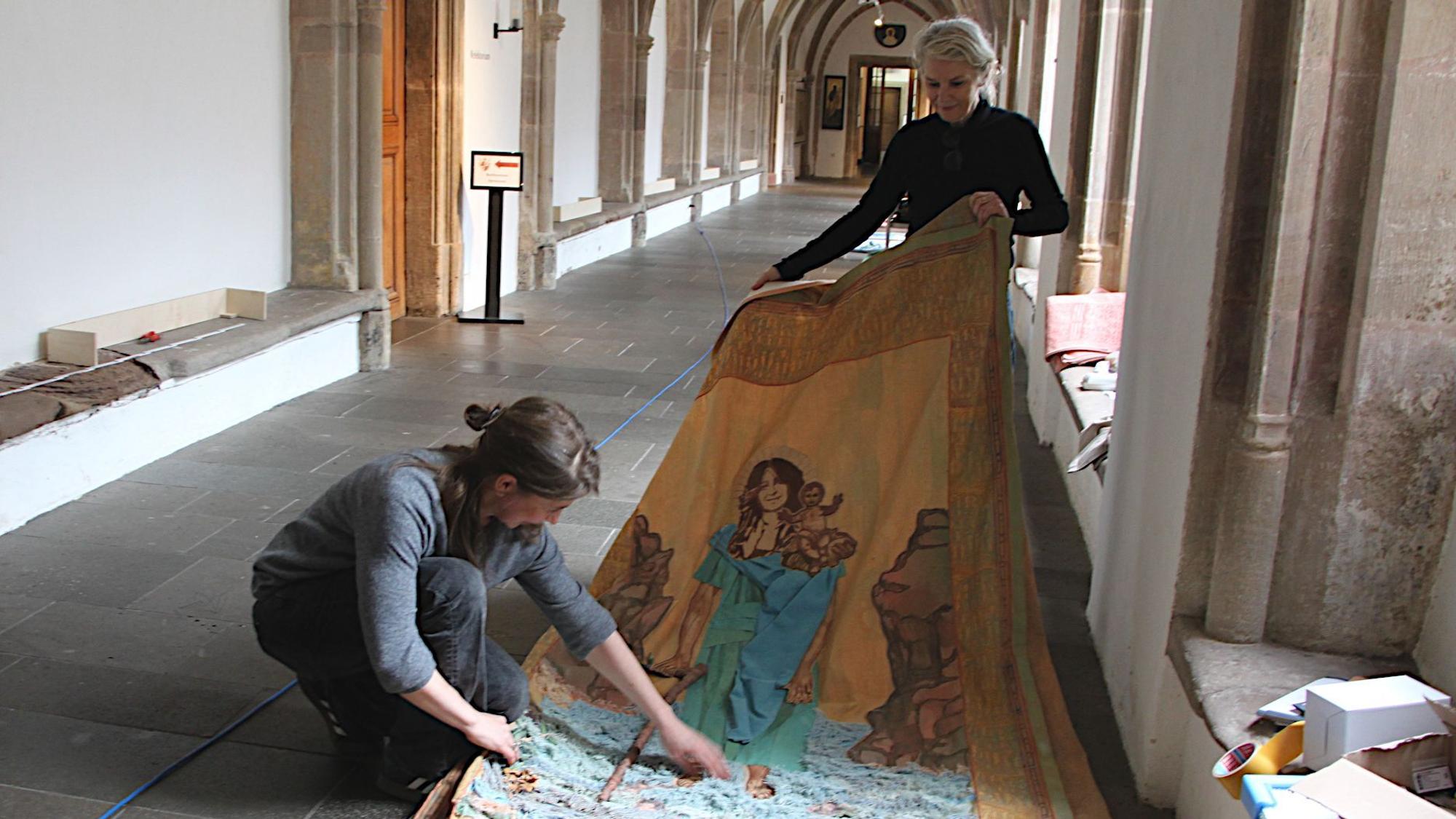 Monika von Bernuth (l.) und Vera Sous halten ein großes Stück Stoff, auf dem Ghada Khano als heiliger Christopherus dargestellt wird. Sie befinden sich im Kreuzgang von Kloster Steinfeld.