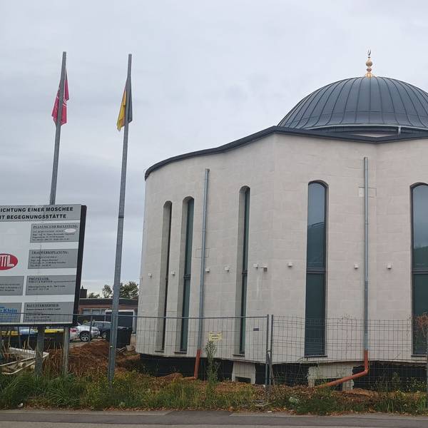 Zu sehen ist das Gebäude der Moschee. Die gebogenen Fenster sind sehr lang und ziehen sich fast über die gesamte Höhe des Gebäudes. Oben ist eine Kuppel.