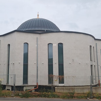 Zu sehen ist das Gebäude der Moschee. Die gebogenen Fenster sind sehr lang und ziehen sich fast über die gesamte Höhe des Gebäudes. Oben ist eine Kuppel.