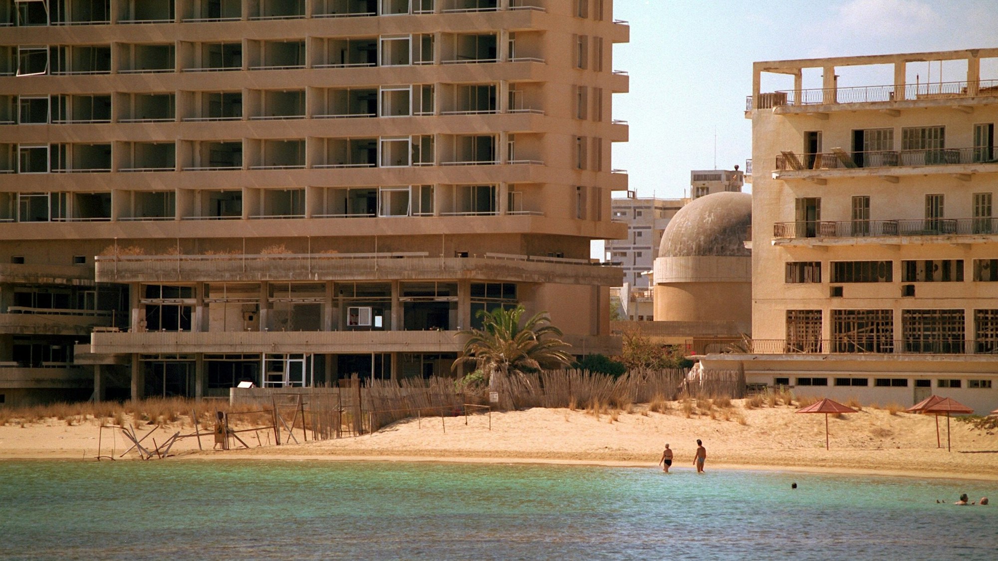 Zwei Touristen waten im Oktober 1998 in Strandnähe durch das flache Wasser. Sie nähern sich dabei der durch einen Stacheldrahtzaun markierten Demarkationslinie, die ihren Hotelkomplex (r) in Gazimagusa (ehemals Famagusta) im Norden Zyperns von den leerstehenden Hotelruinen (l) der früheren Touristenstadt Varosha (Vorort von Famagusta) trennt.