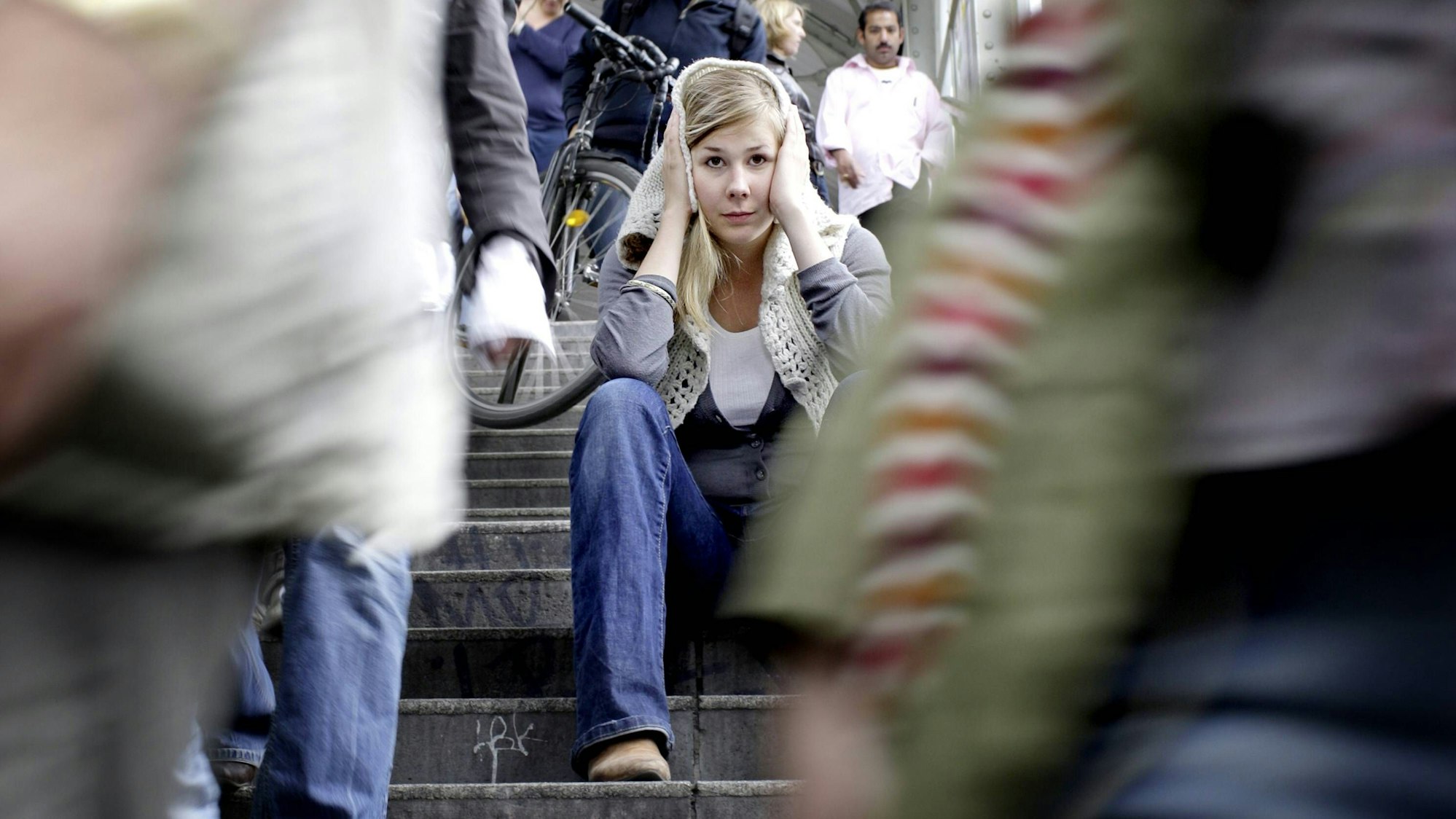 Junge Frau sitzt auf einer Treppe und hält sich die Ohren zu, während um sie herum hektische Menschen unterwegs sind.