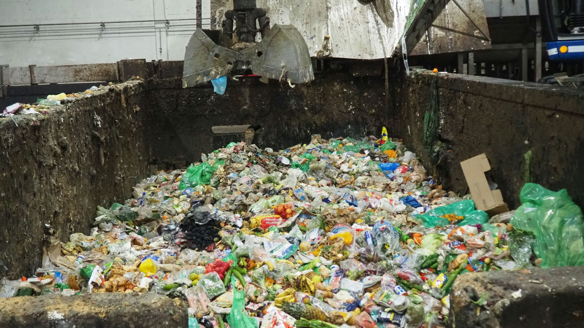 Blick auf den Inhalt eine Betonwanne: Ein geöffneter Baggergreifer schwebt über Verpackungen und Lebensmitteln.