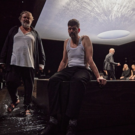 Andreas Becks steht links im Bild, Frank Genser sitzt daneben. Im Hintergrund sieht man den Rest des Ensembles, darüber den mit Wasser gefüllten Baldachin.