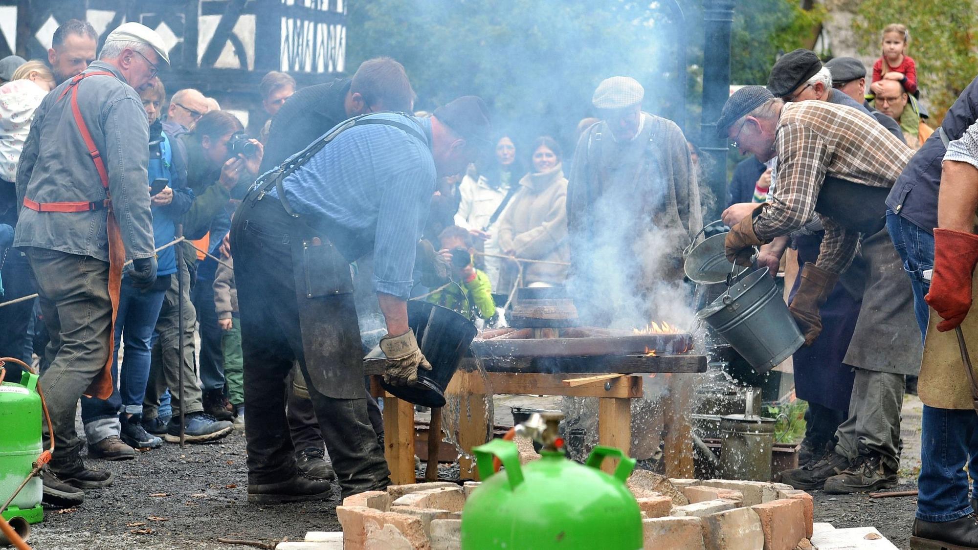 Männer mit Hemden und Schürzen gießen Wasser auf ein Feuer, was Dampf erzeugt.