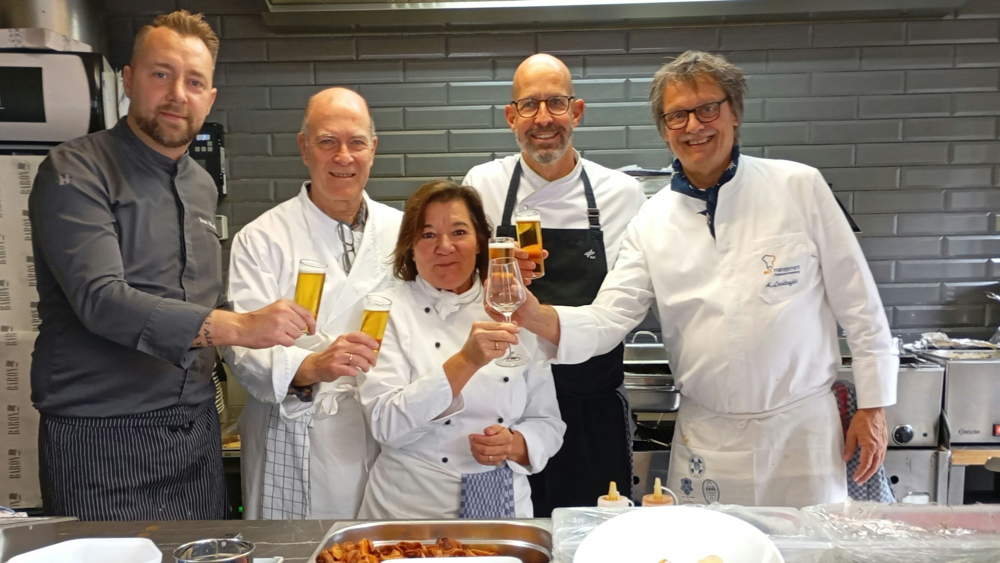 Männer und eine Frau in Kochjacken stehen in einer Küche und stoßen mit Gläsern an.