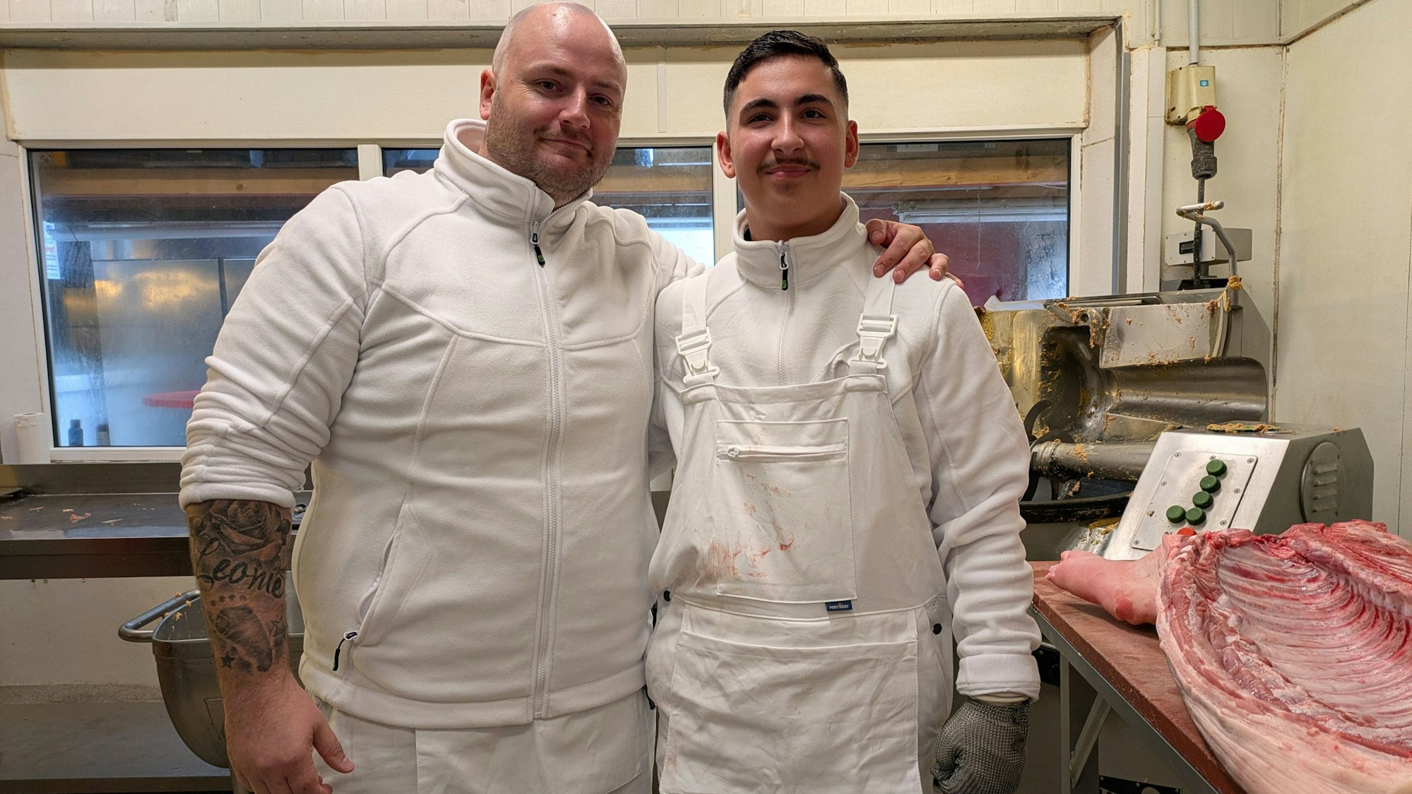 Zwei Männer stehen in einer Fleischerei neben einer Schweinehälfte.