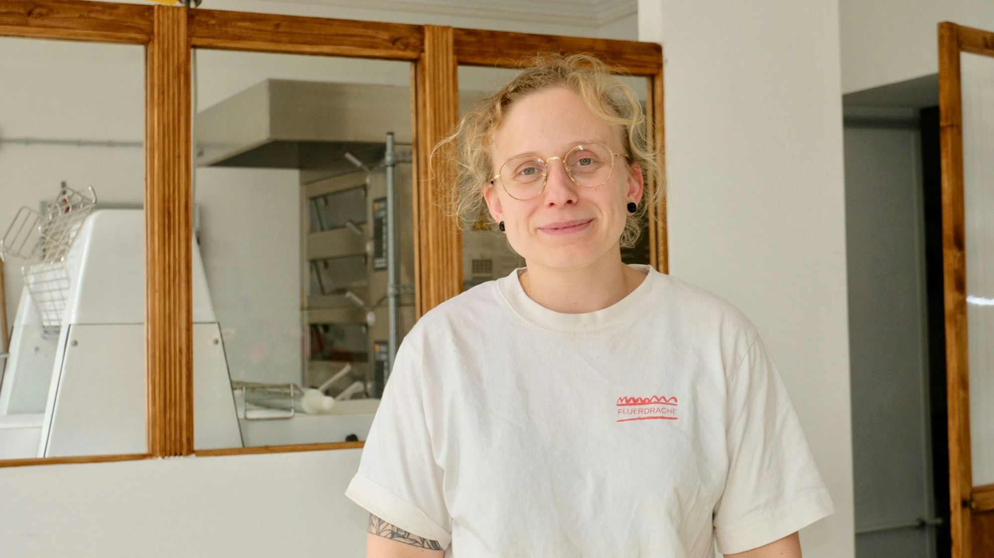 Ein Frau mit blondem Haar und Brille steht vor einer Scheibe, dahinter ist die Backstube zu sehen, in der sie Brot und Croissants backt.
