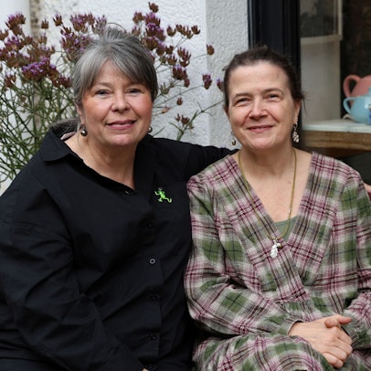 Goldschmiedin Ursula Palm-Zumbé und Künstlerin Annette von der Bey sitzen nebeneinander auf einer Bank vor einem Schaufenster.