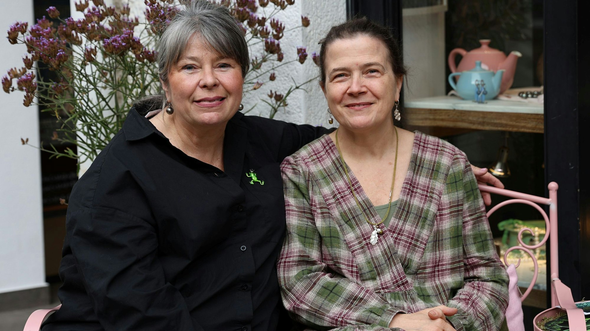 Goldschmiedin Ursula Palm-Zumbé und Künstlerin Annette von der Bey sitzen nebeneinander auf einer Bank vor einem Schaufenster.