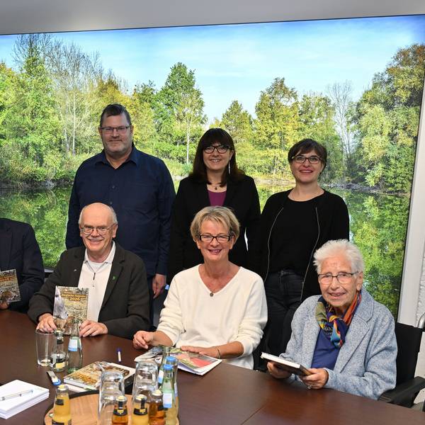 Mehrere Frauen und Männer sitzen an einem Tisch und halten ein Buch in der hand. Hinter ihnen eine grüne Landschaft.
