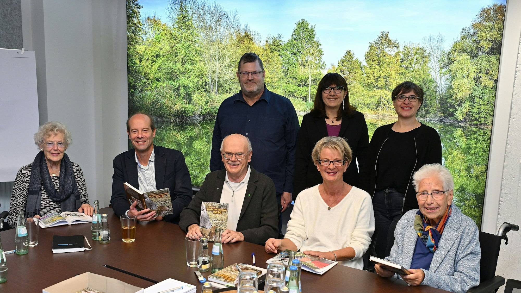 Mehrere Frauen und Männer sitzen an einem Tisch und halten ein Buch in der hand. Hinter ihnen eine grüne Landschaft.