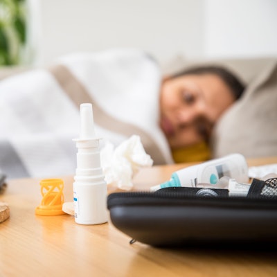 Eine Frau liegt mit einer Decke auf der Couch. Auf dem Tisch vor ihr stehen ein Wasserglas, liegen Taschentücher und Medikamente gegen Erkältung.