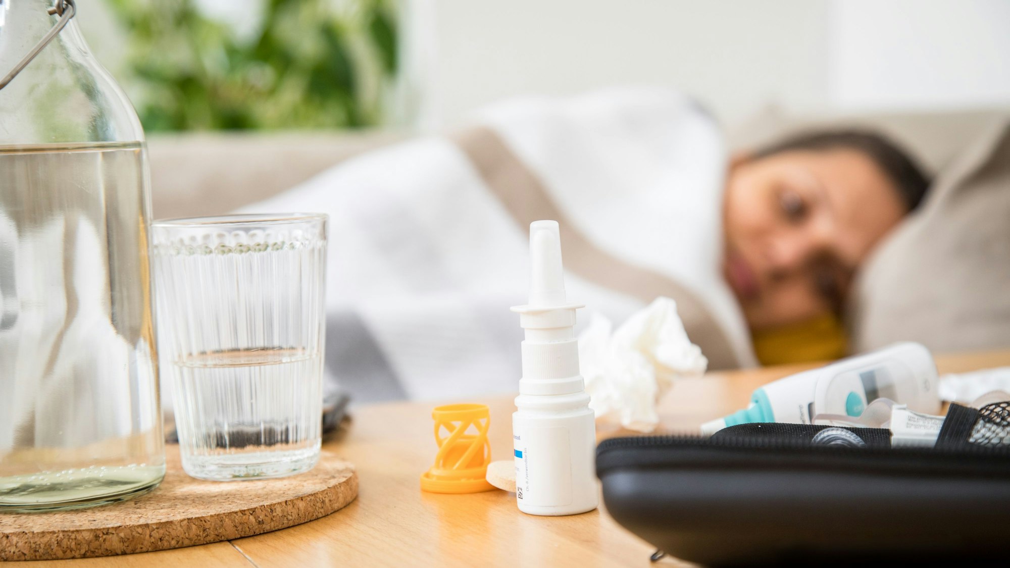 Eine Frau liegt mit einer Decke auf der Couch. Auf dem Tisch vor ihr stehen ein Wasserglas, liegen Taschentücher und Medikamente gegen Erkältung.