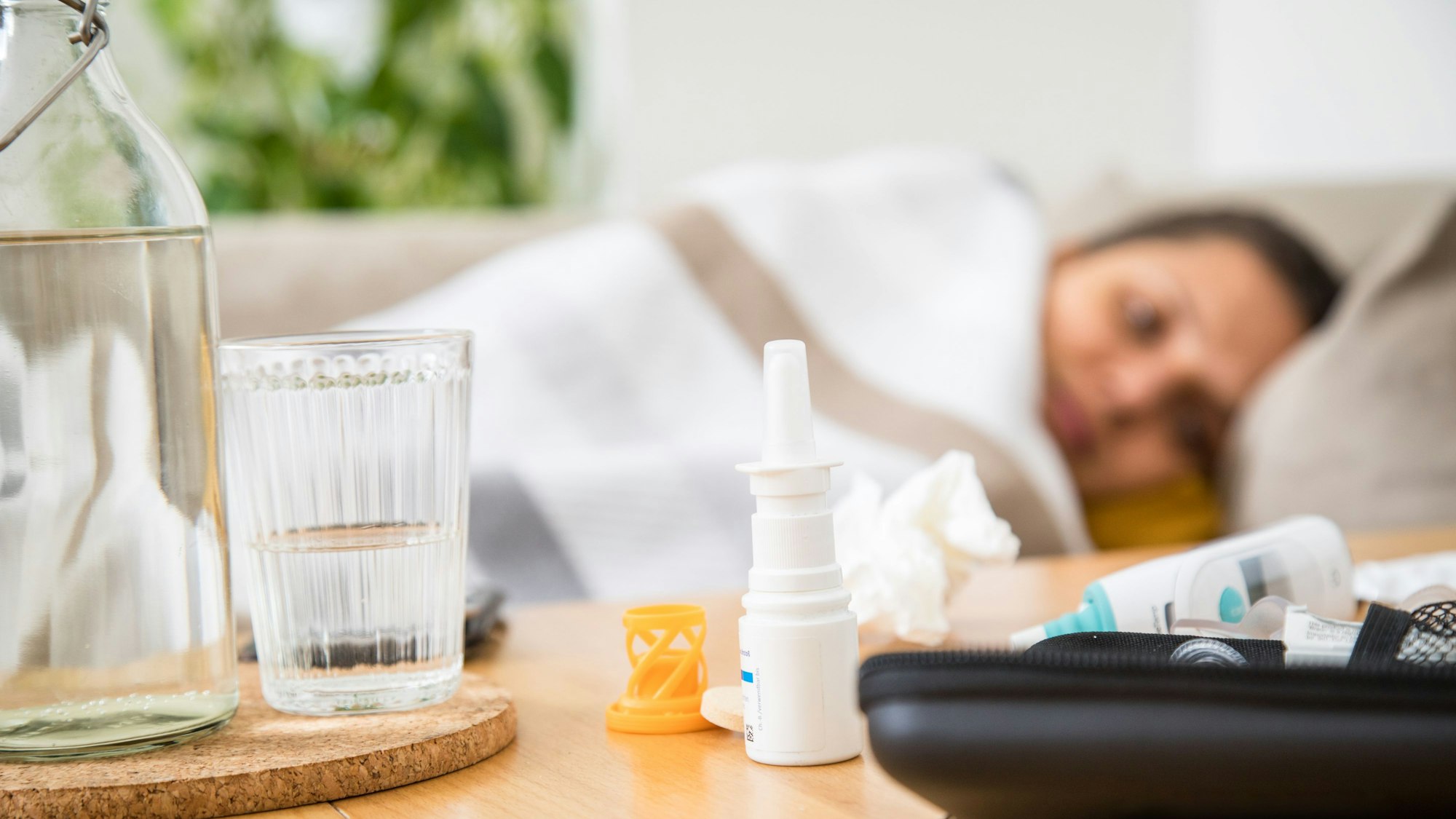 Die Symbolfoto zeigt eine Person, die im Bett liegt, im Vordergrund ein Nasenspray und ein Fieberthermometer.