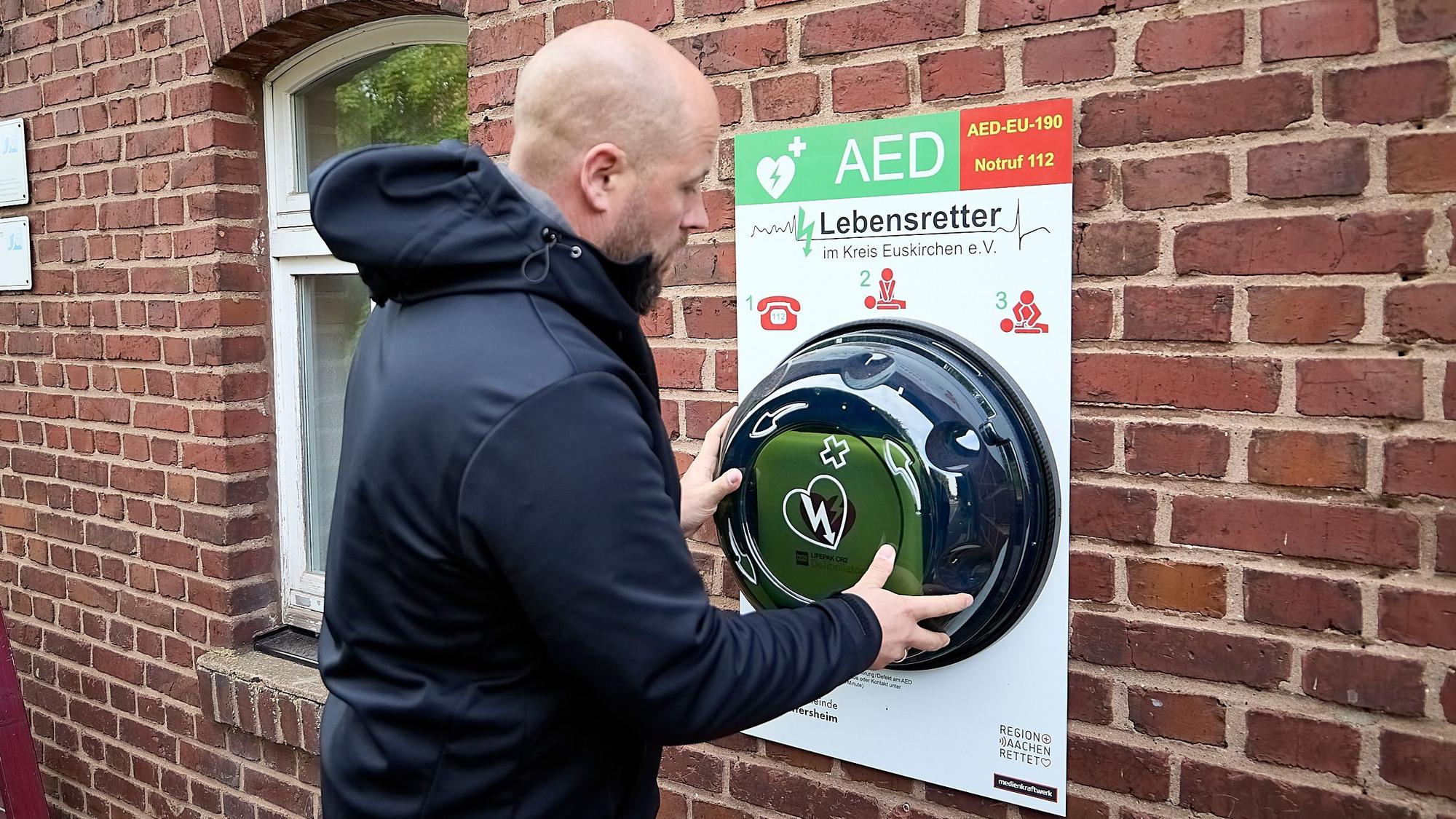 Michael Gissinger steht an einem Defibrillator, der an einer Hauswand installiert ist.
