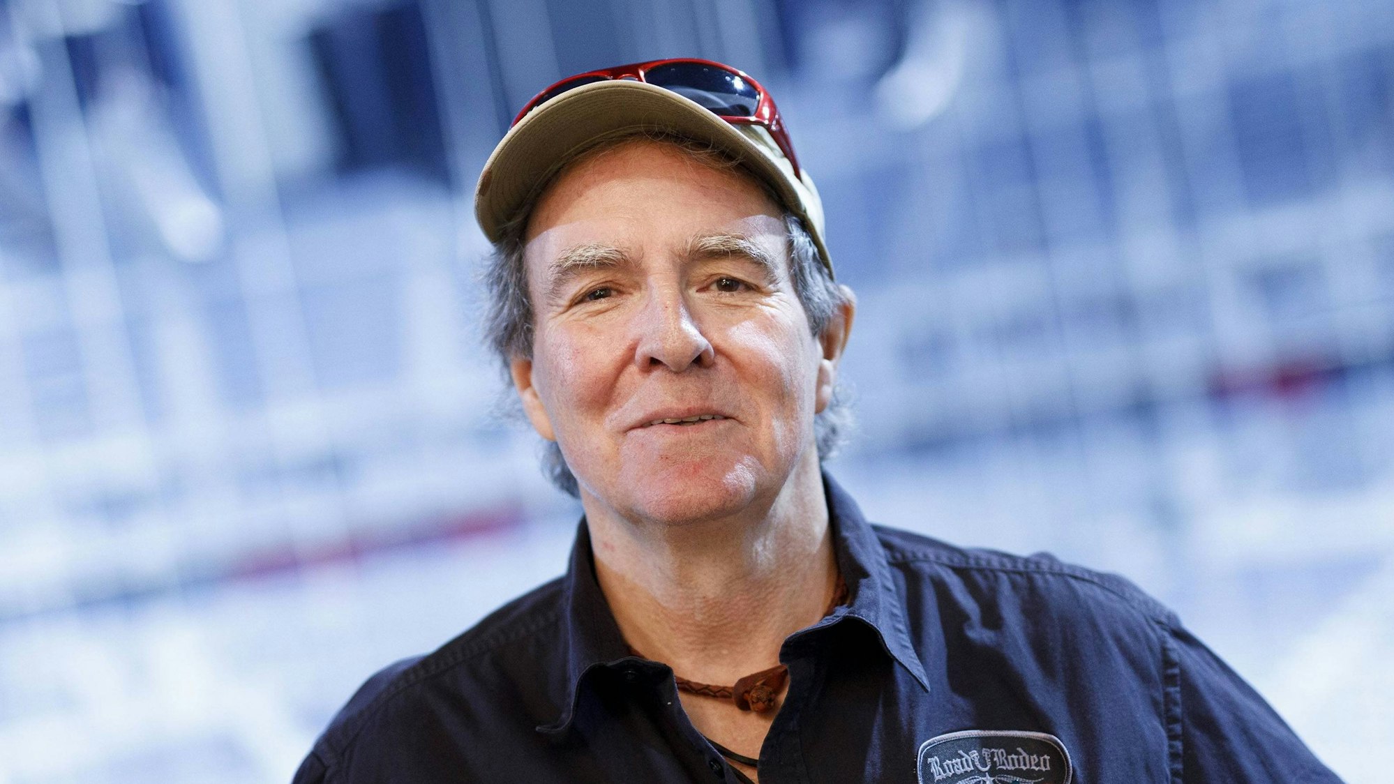 Sänger Peter Horn lächelt mit Kappe, Hemd und „Roadie Rodeo“ Patch vor blauem Hintergrund.