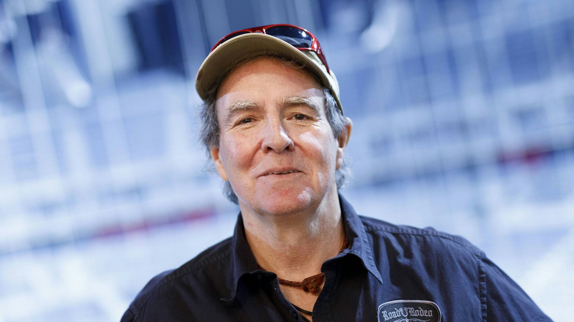 Sänger Peter Horn lächelt mit Kappe, Hemd und „Roadie Rodeo“ Patch vor blauem Hintergrund.